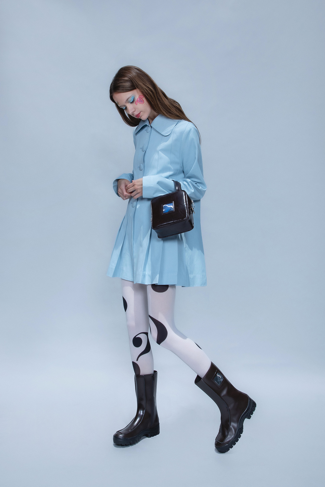 Full body shot of a model in white tights, a blue dress and black boots looking at the ground with hands touching one another against a gray backdrop